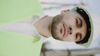 Dışarıdaki genç mühendisin portresi. Ayakta duruyorum, Yapı Arkaplanındaki Kameraya bakıyorum. Kafkas Çekici İnşa Edici veya Güvenlik Giysisi. Dikey Video Giyimi