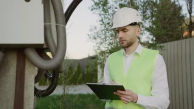 Yakışıklı Erkek Mühendis Sokakta Çalışıyor ve Kalkanı Denetliyor, Not Defterine Not Tutuyor, İş Üniforması Giyiyor. Bakım ve Servis. Güç Hatları Verileri ve Sistemin Çalışmaları Analiz Ediliyor