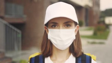 Binanın yanında duran koruyucu maskeli güzel beyaz kadın kuryenin portresi. Hızlı Güvenli Teslimat. Postacı kızın eve teslimatı. Pandemik Kadın Teslimatı. Kapat.