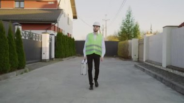 İnşaat işçisi yeni evlere doğru yürüdükten sonra eve dönüyor. Evde tamirat yaptıktan sonra yanında alet çantası taşıyan kasklı bir erkek işçi.