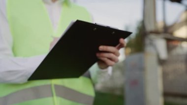 Yelek mühendisinin elleri not alıyor. Tablet kullanarak güç hattını denetliyor. Elinde beyaz kağıt üzerine bir kalemle bir tablet ve notlar tutuyor. Çalışma Planını Analiz Et.