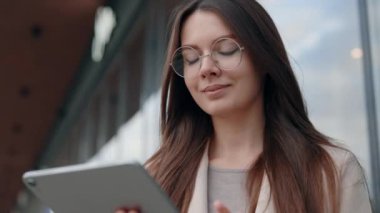 Beyaz tenli çekici iş kadını gözlük takıyor, dijital tablet kullanıyor ve Modern Ofis Merkezinin yanında dururken sosyal medyayı kontrol ediyor. İnsan, Yaşam Tarzı ve Gadget kavramı