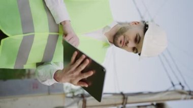 Güç hattı denetimi. Genç Mühendis 'in dikey videosu, Beyaz Şapka ve Yelek Giyiyor, Tabletle İnceleme Yapıyor. Mimarlık Görevindeki Çalışma Planını Analiz Et