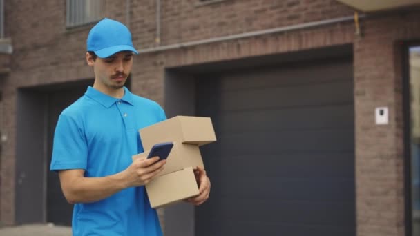 男子快递员手握纸盒和手机沿着大楼走着 派递员在室外派递时使用智能手机在街上寻找派递人地址 — 图库视频影像