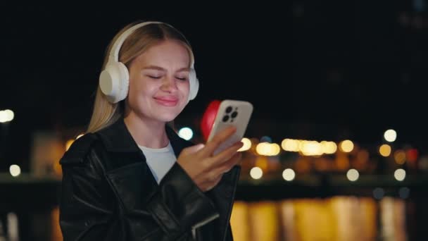 Pretty Girl Usando Teléfono Usando Auriculares Blancos Pie Afuera Noche — Vídeo de stock