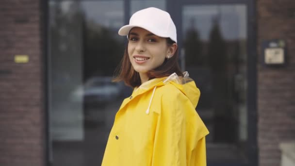 建物の近くに立っているレインコートの笑顔の白人女性クーリエの肖像画 速い配達 特別な黄色のユニフォームとホワイトキャップを着用したポストレディ 労働者 — ストック動画
