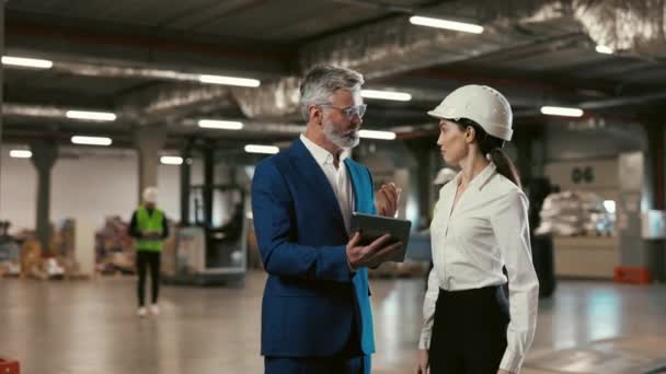 工場のホールに立っている間タブレットと話をする2人のエンジニア タブレットコンピュータを使用しながら新しいプロジェクトを議論するハード帽子の男女産業エンジニア — ストック動画