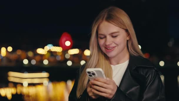 积极金发美女使用智能手机 发短信和微笑 女人在晚上一个人站在外面上网 美丽的女性手持手机 — 图库视频影像