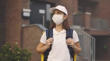 Beyaz Kadın Kurye 'nin Koruma Maskeli Portresi. Binanın yanında duruyor. Hızlı Güvenli Teslimat. Posta Kadını 'nın eve teslimatı kameraya bakıyor. Pandemik Kadın Teslimatı