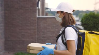 Beyaz kadın kuryenin müşteri evine gidişinin, pizzayı tutuşunun, koruyucu maskeyi takışının, salgın sırasında fast food dağıtımının yan görüntüsü. Yemek getiren kadın Intercom 'u arıyor.
