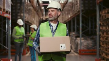 Hard Hat Holding Karton Kutusu Giyen, Ürünler Organize Raflarıyla Dolu Perakende Depo Alanında Yürüyen Kendine Güvenen İşçi. Hızlı tempolu Taşıma ve Dağıtım Merkezi.