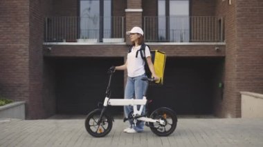 Ofis binasının yanında duran güzel gülümseyen kız kuryenin portresi. Bisiklet Teslimatı. Postacı kadın özel beyaz üniforma giyiyor, sarı sırt çantası ve şapka taşıyor.