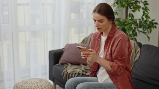 Jovem Caucasiana Sentada Sofá Usando Smartphone Lendo Boas Novas Fazendo — Vídeo de Stock