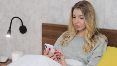 Sarışın, tatlı bir kadın otelde ya da evde yatakta oturmuş arkadaşlarıyla sohbet ediyor ya da akıllı telefondan sosyal ağlara kayıyor. İnsanlar ve teknoloji kavramı. Kapat.