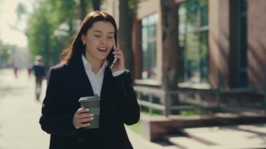 Resmi takım elbiseli mutlu iş kadını iş merkezinin yakınına gidiyor ve müşterileri elinde kahve fincanıyla telefonda konuşuyor. Dışarı çıkın. İnsanlar ve teknoloji kavramı