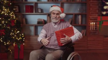 Tekerlekli sandalyede oturan, Noel Baba Şapkası takan, yeni yıl hediyesini tutan ve kameraya başparmağını gösteren yaşlı adamın portresi. Yeni Yıl ve Engelliler Konsepti