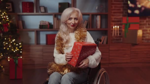 クリスマスプレゼントを持っている車椅子で幸せな女性 メリークリスマスかハッピーニューイヤー グリーンクリスマスツリーの背景に表示されている障害者の笑顔の女性 障害者コンセプト — ストック動画
