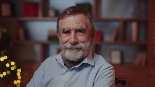 Noel süslemeleriyle dolu odada, tekerlekli sandalyede oturan ve kameraya bakan kıdemli bir adam. Engelli Yaşlı Erkek, Yalnız Büyükbaba Kış Tatillerini Kutluyor. Yeni Yıl Konsepti