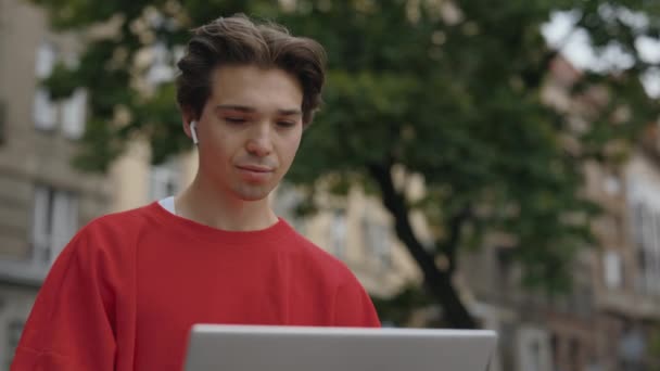 坐在外面的笔记本电脑上 戴着耳机打字的家伙 外面的白人工作隐约 无忧无虑的男人休息和使用笔记本电脑 概念教育 — 图库视频影像