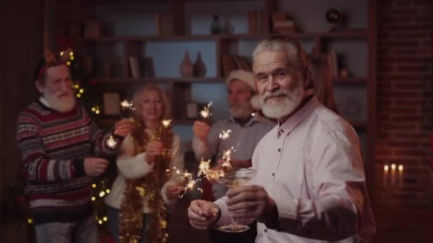 ハンサム シニア スタンディングの肖像画 グラス スパークル ワインを持ち 友人たちがバックグラウンドで踊っている 新年パーティー 一緒に祝い そして笑う — ストック動画