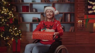 Tekerlekli sandalyedeki gülümseyen yaşlı kadın kameraya yeni yıl hediyesini gösteriyor ve tutuyor. Dekorasyon Evi 'nde oturan engelli kadın Noel Hediyesi veriyor. Engelliler ve Tatil Konsepti