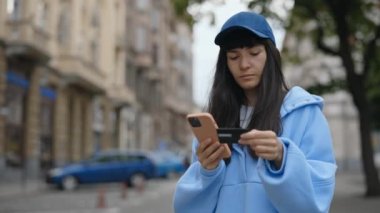 Beyaz bir kadın, dışarıda dururken online alışveriş için kredi kartıyla ödeme yapıyor. Sokakta Telefonu Kullanan Gülümseyen Kadın. Kredi Kartı Tutarken Cep Telefonu Kullanan Kadın