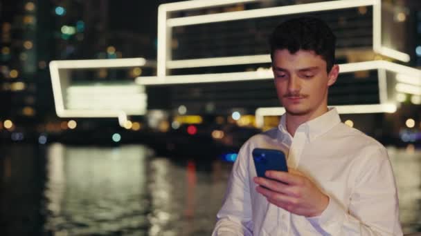 盖伊游客站在堤岸背景下使用智能手机 千禧年男子在夜城街的屏幕上从事翻书和打字工作 男性在移动设备上浏览互联网 — 图库视频影像