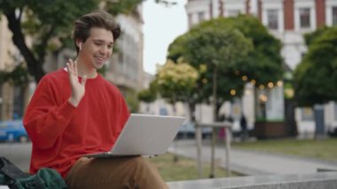 Günlük kıyafetlerle gezen neşeli genç bir adam dışarıda bir video görüşmesi yapıyor, dizüstü bilgisayar kullanırken hareketli hareketler yapıyor. Halk iletişimi