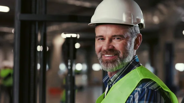 Güvenli Yelek ve Sert Şapkalı Gülümseyen Olgun İşçi Portresi. Forklift sürücüsü kameraya bakıyor. Teslimat Mallarıyla dolu raflı bir depo. Dağıtım Lojistik Merkezi ve Halk Kavramı