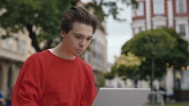 Heyecanlı Genç Yetişkin Adam dışarıda bankta otururken dizüstü bilgisayara bakarak zaferini kutluyor. Günlük giysiler içindeki yakışıklı beyaz adam iyi haberler okuyor, kazanmaktan dolayı mutlu.