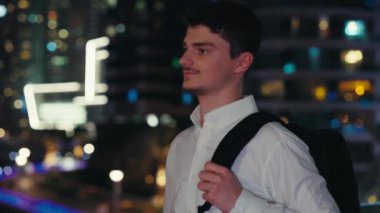 Akşamları dışarıda yürüyen beyaz, yakışıklı, erkek turist. Genç Gezgin Sırt çantasıyla Modern Gece Şehrinin Sokakları 'nı Keşfediyor. Gece Gezileri ve İnsanlar Konsepti