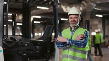 Güvenlik Üniforması Giyen İşçi, Ayakta Duran, Gülümseyen, Kollarını çaprazlayan ve Forklift yakınlarındaki bir fabrikada Mutlu Olmak. Profesyonel Endüstri 'de çalışan yakın çekim adamları. Lojistik Merkezi