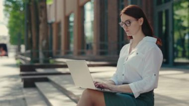 Resmi takım elbiseli, gülen kızıl saçlı iş kadını iş merkezinin merdivenlerinde oturmuş laptopla çalışıyor. İnsanlar ve teknoloji kavramı