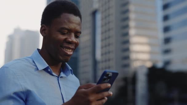 ハッピーアフリカ系アメリカ人のマネージャーが外に立ち 電話でインターネットを閲覧しています 笑顔ビジネスマン モバイルでビデオを見ている屋外 人と技術のコンセプト — ストック動画