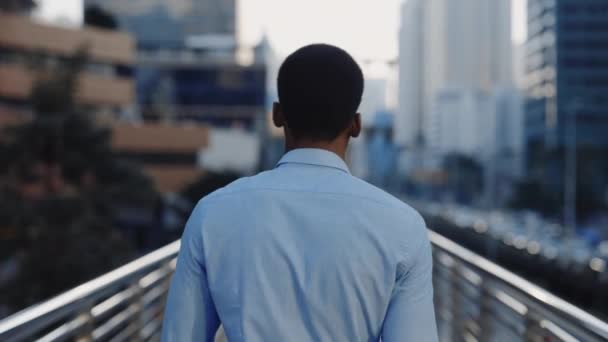 ビジネス地区のクロスブリッジを歩くシャツで成功した若いアフリカ系アメリカ人男性のバックビュー 仕事への道を歩くビジネスマンの背中 人々のコンセプト — ストック動画