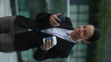 Dikey video. Çekici iş kadını dışarıda kahve molasında akıllı telefon kullanarak videolar izliyor. Gülümseyen Etnik Bayan Lider Dışarıda Çay İçen, Cep Telefonu Tutan, İnsan ve Teknoloji Konsepti