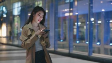 Şık Cinsiyet Değiştiren Kadın Şehirde Yürüyor ve Telefonu Kullanıyor. Asyalı Genç Kadın Gece Sokakta Aşağı Gidiyor ve Arkadaşlarıyla Sohbet Ediyor. Halk ve Teknoloji Konsepti