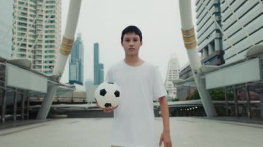 Asyalı çocuk elinde futbol topunu tutarken dışarıda kameraya bakıyor. Şehrin arka planındaki çok ırklı genç futbolcunun portresi. Halk ve Futbol Konsepti