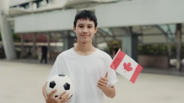 Asya Gençleri Futbol Hayranı 'nın Kanada Bayrağı ve Balosu ile Açık Hava Poz Verme portresi. Futbol topu ve Kanada bayrağıyla dışarıda duran genç adam. Futbol ve İnsanlar Konsepti