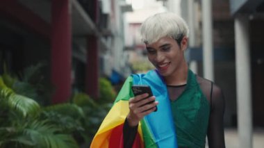 Transseksüel adam Lgbt bayrağına sarılmış, sokak kaydırma uygulamalarına gidiyor. İnternetteki Yakışıklı Modern Homoseksüel Dış Tarayıcı Cep Telefonu Kullanımı. LGBT ve Teknoloji Konsepti