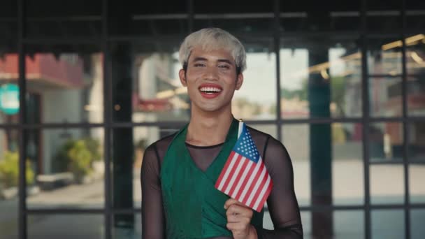 メイクアップとカメラを見ながら立っている男性愛国者 トランスジェンダーのハンサムな若い男の外側に立って米国の旗を保持する肖像画 ダイバーシティコンセプト — ストック動画