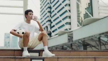 Asyalı erkek sporcu, uzun bir spor antrenmanından sonra dışarıda elinde topla içki içiyor. Genç futbolcu susamış hissederek su içiyor. Futbol ve İnsanlar Konsepti