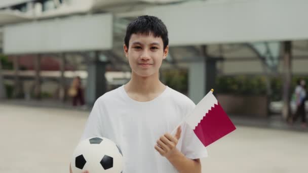 アジアの少年がカタールの旗を掲げ スタジアムの背景にあるサッカーボールと立って カメラを見ている カタールの旗とサッカーボールとの試合前に若いスポーツファン サッカーと人々のコンセプト — ストック動画