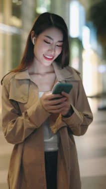 Dikey video. Yakışıklı Asyalı İş Kızı Cep Telefonu Tutuyor, Arkadaşlarıyla Sohbet Ediyor. Sosyal Uygulamada Akıllı Telefon Mesajı Kullanan Kadın Dışarıda Dururken. Halk ve Teknoloji Konsepti