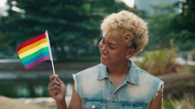 Parkta Küçük bir LGBT Gökkuşağı Bayrağı Tutan Gülümseyen Genç Adam, Çeşitlilik ve Gururu Kutluyor. Denim yeleği giyip Neşe ve Ayrıcalık saçar..