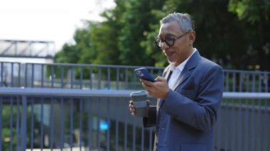 Gülümseyen Asyalı Neşeli Adam Köprüde yürüyor, elinde Eko Kupa var ve Akıllı Telefonu kullanıyor. Kıdemli iş adamı kahve molasından dönüyor ve telefonda komik videolar izliyor..