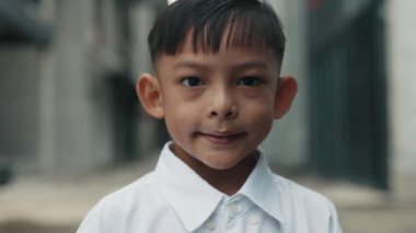 Beyaz gömlekli küçük Asyalı çocuk şehir caddesinde duruyor. Okul üniforması giyen okul çocuğunun portresi çok katlı binalarla birlikte güvenli bir şekilde mahallede yürüyor.