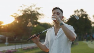 Akşam portresi için susamış bir adam maç üstüne tatlı su içiyor. Golf sopasıyla yorgun sporcu, Alacakaranlık Parkı 'nda içkiyle serinliyor. Turnuvada sıvı şişeli erkek golfçü