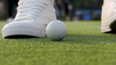 Golfçü küçük topa vurmaya geliyor. Parlak sürücüsü ile birlikte çimlerin yemyeşil olduğu yerde. Beyaz ayakkabılı sporcu parkta golf oynar. Banliyö kulübünde rekabetçi bir spor oyunu