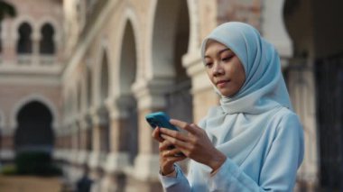 Bir binanın önünde tesettürlü genç bir Asyalı kız arkadaşlarıyla cep telefonunda sohbet ediyor, kameraya Müslüman Kadın Pozları çekiyor, akıllı telefonuyla mesaj yazıyor. Halk ve Teknoloji
